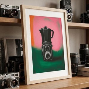 May include: A framed art print featuring a black moka pot coffee maker combined with a twin-lens reflex camera body. The illustration is set against a vibrant background of blended pink, orange, and deep green colors in a light wood frame.