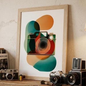 May include: A framed wall art print featuring a stylized camera illustration overlaid with abstract, overlapping shapes in shades of orange, teal, and red. The camera is depicted with clean lines, centered against a white background within a light wood frame.