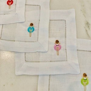 May include: Four white linen napkins with embroidered images of a person in a swimsuit floating on a colorful inflatable ring. The rings are red, blue, pink, and green.