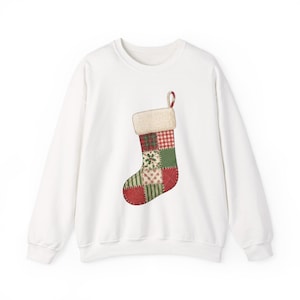 May include: A white long-sleeve sweatshirt featuring a central graphic of a Christmas stocking. The stocking is designed with a patchwork pattern in red, green, and white, including plaid, gingham, and floral prints, topped with a fuzzy white cuff.
