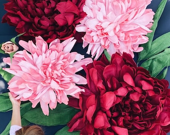 Könnte beinhalten: Eine Wanddekoration mit fünf großen Papier-Pfingstrosen in Rosatönen und Burgunderrot. Die Blumen sind in einer Gruppe mit grünen Blättern angeordnet und vor einem dunkelblauen Hintergrund platziert.