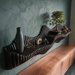 May include: A dark brown, undulating, slatted wall-mounted shelf featuring a series of vertical wooden fins. The shelf holds a tall gray vase, a gold geometric wire sculpture, two polished stone specimens, a small bonsai tree, and dried pods.