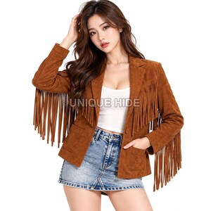 May include: A brown suede blazer featuring long fringe detailing along the chest, shoulders, and sleeves. The jacket includes a single button closure, two front pockets, and a notched lapel. The text UNIQUE HIDE is printed on the front.