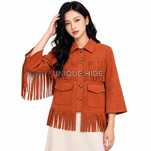 May include: A burnt orange jacket featuring long fringe detailing on the sleeves and hem, a collared neckline, button-front closure, and two front flap pockets. The text UNIQUE HIDE is printed across the chest area of the garment.