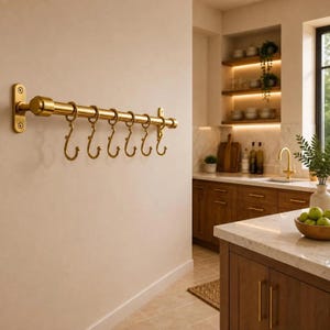 May include: A wall-mounted gold-toned metal rail featuring six matching S-shaped hooks suspended from circular rings. The horizontal bar is secured to the wall with two flat, rectangular brackets, each fastened with visible screws at both ends.
