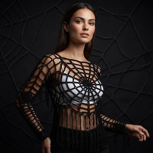 May include: A black long-sleeved crochet top featuring an intricate spider web pattern across the chest and sleeves, finished with a fringed hemline. The garment is worn over a solid white base layer, showcasing the open-knit texture and design.