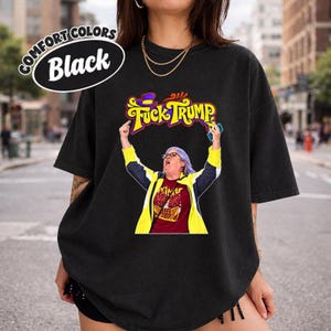 May include: A black t-shirt featuring a graphic of a person with short hair and glasses, wearing a maroon shirt under a yellow and navy jacket. The person has both arms raised, and the text "FUCK TRUMP" is printed in yellow above them.