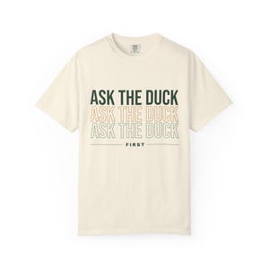 May include: A cream-colored short-sleeve t-shirt featuring the text ASK THE DUCK repeated three times in stacked lines with varying font styles and colors, followed by the word FIRST underlined by a horizontal line centered on the chest.