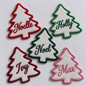 May include: Five white fabric Christmas tree patches with embroidered borders and names. The patches feature the names Noelle in red, Holly in green, Noel in green, Ivy in dark red, and Max in pink, each stitched in a cursive font.