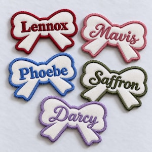 May include: Five white fabric bow-shaped patches with embroidered borders and names. The names are Lennox in red, Mavis in pink, Phoebe in blue, Saffron in olive green, and Darcy in purple, all stitched in a script or block font.