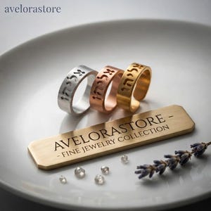 Puede incluir: Tres anillos de metal en tonos plata, cobre y oro, cada uno con caracteres hebreos grabados. Una placa rectangular de metal dorado muestra el texto AVELORASTORE - FINE JEWELRY COLLECTION. Se incluyen pequeñas piedras transparentes y una rama de lavanda.