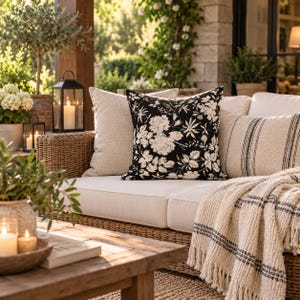 Peut inclure: Un coussin carré avec un fond noir et un motif floral blanc, placé sur un canapé en osier aux côtés d'un coussin beige uni, d'un coussin lombaire rayé et d'un plaid crème à franges disposé sur l'assise.