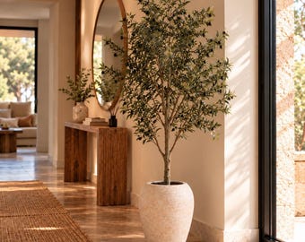 May include: An artificial olive tree with green leaves and small dark olives, planted in a large, textured, light-colored stone-like planter. The tree has a central trunk that branches out into a full canopy of foliage.