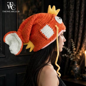 May include: An orange crochet hat shaped like a fish with a large tail, white side patches, yellow fins on top and sides, and long yellow whiskers. The hat features large white eyes with black pupils and a light pink trim along the bottom edge.