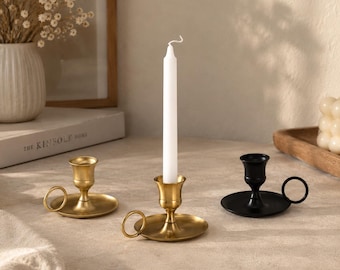May include: Three metal candle holders with circular bases and side ring handles. Two holders have a brushed gold finish, and one holder has a matte black finish. A single white taper candle is inserted into the centre gold candle holder.