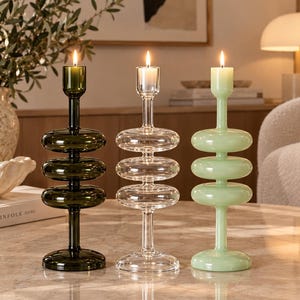 May include: Three glass candle holders stand in a row, each featuring a central stem with three stacked, rounded, disc-like shapes. The left holder is dark olive green, the center is clear, and the right is a pale mint green, all holding lit candles.
