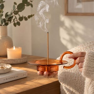 Può includere: Un porta incenso in vetro color ambra con base circolare e manico laterale curvo. Un singolo bastoncino di incenso sottile è posizionato verticalmente al centro del supporto, emettendo una scia visibile di fumo bianco che sale sopra il vetro.