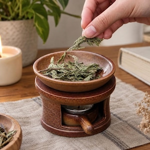 May include: A brown ceramic oil burner with a textured finish, featuring a removable top dish filled with dried green herbs. The base has a hollow center containing a small tea light candle holder with a handle, designed for heating.