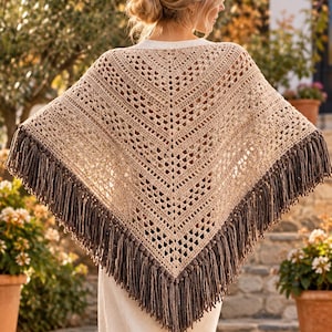 May include: A beige crochet poncho featuring a V-shaped design with an intricate openwork pattern. The garment is finished with a long, dark brown fringe along the bottom edges, creating a textured and layered appearance over a white top.