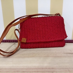 Peut inclure: Sac bandoulière rouge en crochet avec fermeture à rabat et une bandoulière en cuir marron. Le sac porte une petite étiquette rectangulaire avec le mot "LOVE". Le sac est posé sur une surface en bois.