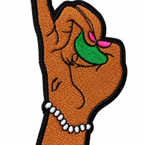 May include: An embroidered patch featuring a brown hand making a gesture with the index finger pointing upward. The hand wears a white beaded bracelet and holds a bright green crescent shape with two small pink accents, all outlined in black.