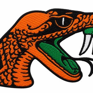 May include: An embroidered patch featuring the head of a snake in profile. The snake has bright orange scales with black outlines, a wide-open mouth with a green interior, a black forked tongue, and two prominent white fangs with black borders.