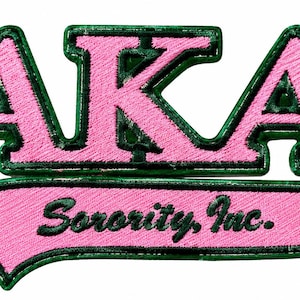 May include: An embroidered patch featuring the letters AKA in pink with a dark green border. Below the letters, a pink swoosh shape contains the black embroidered text Sorority, Inc. The entire design is outlined in dark green thread.