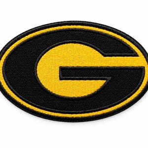 May include: An oval-shaped embroidered patch featuring a bold yellow letter G centered on a black background. The patch has a thick black border with a thin yellow inner ring, creating a high-contrast design with visible stitching texture.