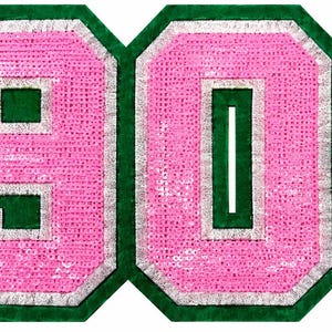 以下が含まれることがあります： カレッジフォントで数字の1908がデザインされた大きな装飾用ワッペン。数字は鮮やかなピンクのスパンコールで埋め尽くされ、シルバーの縁取りが施されており、ダークグリーンのフェルト生地の背景にグリーンの縁取りが組み合わされています。
