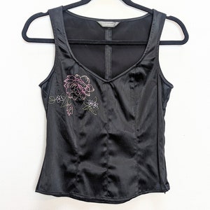 May include: Black satin tank top with a V-neck and floral embroidery in pink and green. The top is hanging on a black hanger against a white background. The top has a fitted silhouette and a subtle sheen.