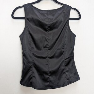 May include: A sleeveless black satin top hangs on a black hanger against a white background. The top has a scoop neckline and a fitted silhouette with vertical seams. The fabric has a subtle sheen.