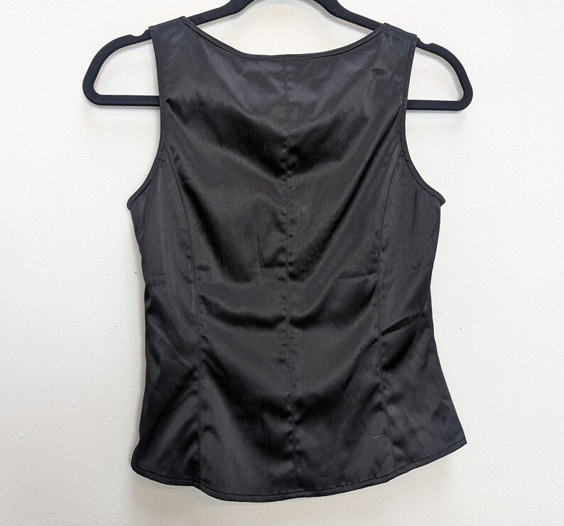 May include: A sleeveless black satin top hangs on a black hanger against a white background. The top has a scoop neckline and a fitted silhouette with vertical seams. The fabric has a subtle sheen.