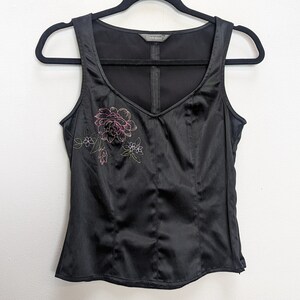 May include: Black satin tank top with a V-neck and embroidered floral design in pink and green. The top is sleeveless and hangs on a black hanger against a white background. The top has a fitted silhouette.