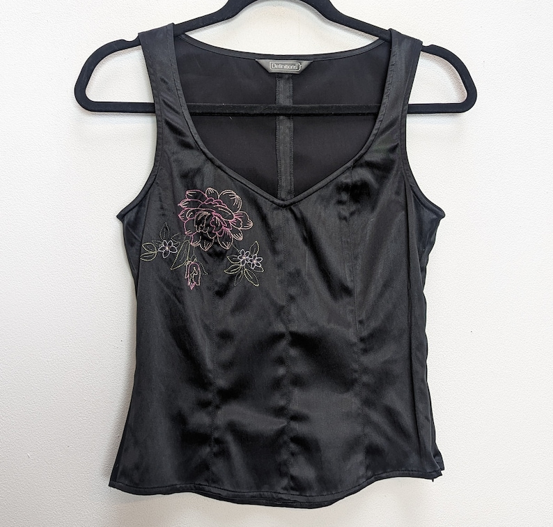 May include: Black satin tank top with a V-neck and embroidered floral design in pink and green. The top is sleeveless and hangs on a black hanger against a white background. The top has a fitted silhouette.
