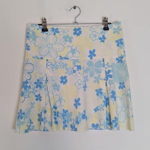 Könnte beinhalten: Weißer Minirock mit einem Blumenmuster in Hellblau und Gelb. Der Rock ist plissiert und hängt an einem Metallbügel. Das florale Design zeigt Blumen in verschiedenen Größen.