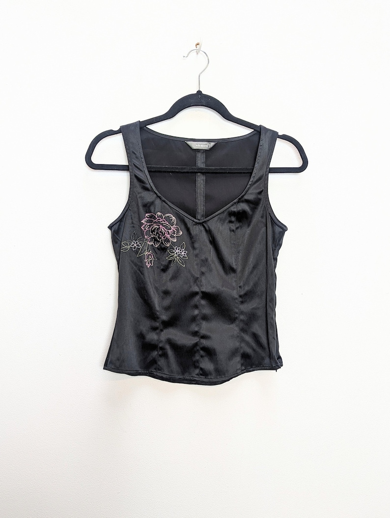 May include: Black satin tank top with a V-neck and floral embroidery in pink and green. The top is hanging on a black hanger against a white background. The top has a fitted silhouette and a subtle sheen.