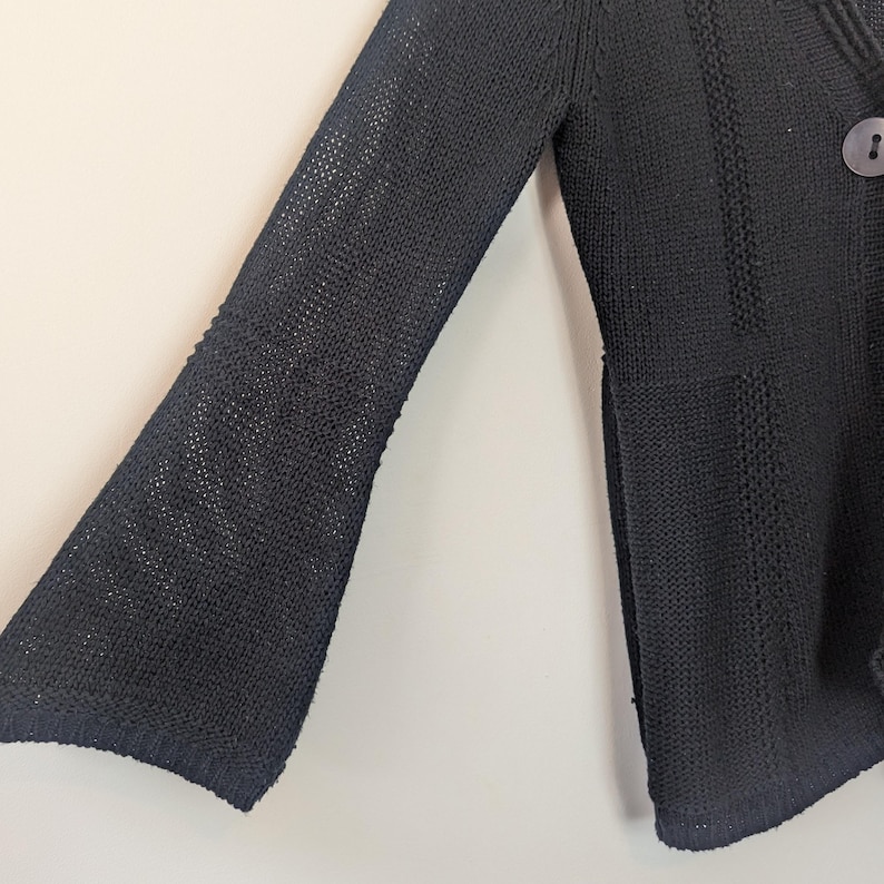May include: Black knitted cardigan with long sleeves and a bell-shaped sleeve design. The cardigan has a single button closure at the neckline.