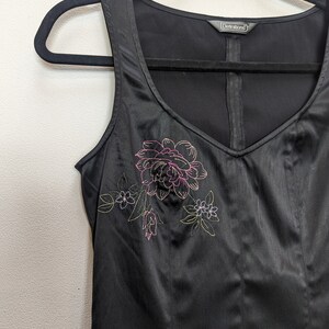 May include: Black sleeveless top with a V-neck and floral embroidery. The embroidered design features a large pink flower with green and yellow accents. The top is hanging on a black hanger.