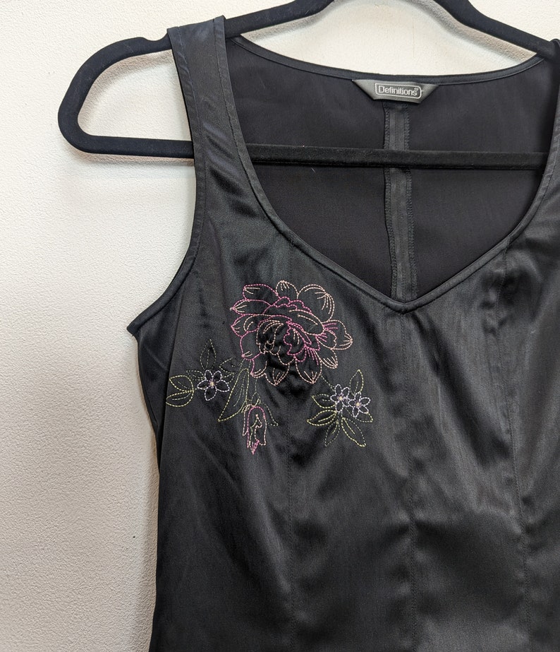 May include: Black sleeveless top with a V-neck and floral embroidery. The embroidered design features a large pink flower with green and yellow accents. The top is hanging on a black hanger.