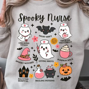 May include: A light gray sweatshirt with a Halloween-themed design. The design includes the text "Spooky Nurse" and various illustrations of ghosts, bats, a witch's hat, a pumpkin, and other Halloween-related elements.