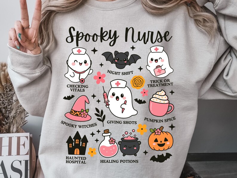 May include: A light gray sweatshirt with a Halloween-themed design. The design includes the text "Spooky Nurse" and various illustrations of ghosts, bats, a witch's hat, a pumpkin, and other Halloween-related elements.