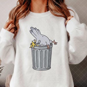 May include: A white long-sleeve sweatshirt featuring a graphic illustration of a grey pigeon diving headfirst into a grey metal trash can. The trash can contains various pieces of refuse, including a yellow banana peel and a small metal tin.