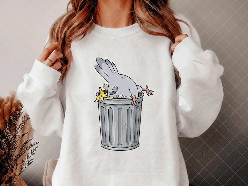 May include: A white long-sleeve sweatshirt featuring a graphic illustration of a grey pigeon diving headfirst into a grey metal trash can. The trash can contains various pieces of refuse, including a yellow banana peel and a small metal tin.