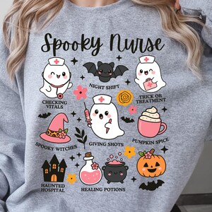 May include: A light gray sweatshirt with the words "Spooky Nurse" in a script font. The design features cartoon ghosts, bats, and Halloween-themed icons like a witch's hat and pumpkin. The phrases "Checking Vitals" and "Pumpkin Spice" are included.
