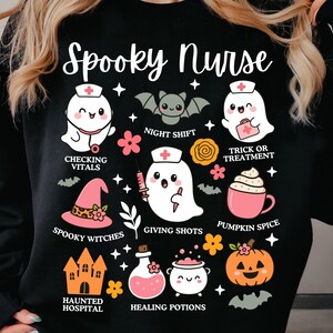 May include: Black sweatshirt with a "Spooky Nurse" graphic. The design features cartoon ghosts, bats, and Halloween-themed icons like a witch's hat, pumpkin, and potion bottles. Text includes "Checking Vitals" and "Giving Shots."