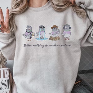May include: A beige crewneck sweatshirt featuring a graphic of four cartoon pigeons in various states of distress, with the text "Relax, nothing is under control" printed in black cursive script underneath the illustrations of the birds.
