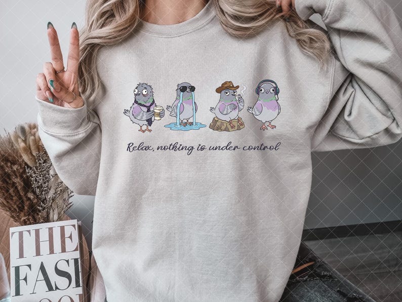 May include: A beige crewneck sweatshirt featuring a graphic of four cartoon pigeons in various states of distress, with the text "Relax, nothing is under control" printed in black cursive script underneath the illustrations of the birds.