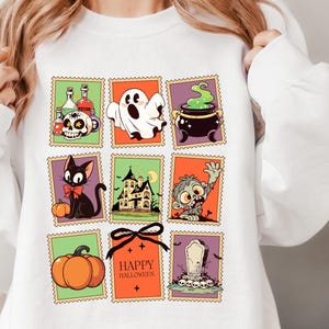 May include: White sweatshirt featuring a colorful Halloween-themed design. The design includes various illustrations such as a skull, ghost, cauldron, black cat, haunted house, zombie, pumpkin, and the text "Happy Halloween".