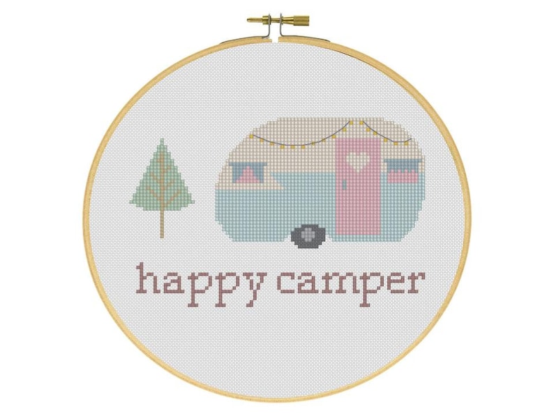 May include: Cross-stitch embroidery in a wooden hoop features a light blue and pink camper with a string of lights, a green tree, and the words "happy camper" in brown. The design is on white fabric.