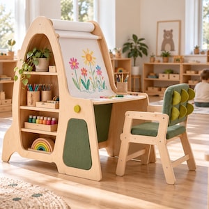 May include: A light-colored wooden children's art desk with an integrated paper roll, side shelving, and a matching chair. The desk features a drawing surface with a floral illustration, while the chair has a green upholstered seat and backrest.